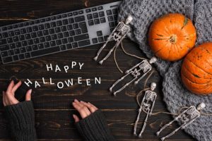 Female hands and Halloween decorations with skeleton and laptop computer on dark wooden desk table. Top view, flat lay. Festival background concept
